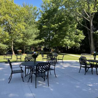 An outdoor patio area with tables and chairs under a sunny blue sky, surrounded by lush green trees.