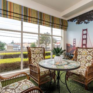 A cozy indoor seating area with a round glass table and patterned chairs, set against a window and a wall mural of the Golden Gate Bridge.