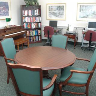 A meeting room with a round wooden table, green upholstered chairs, a bookshelf filled with books, two computers on desks, a piano, and framed artwork on the walls.