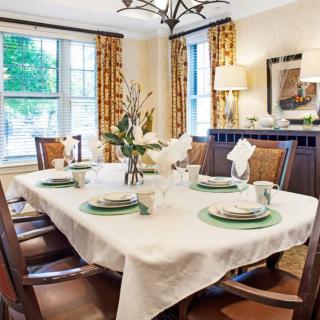 An elegantly set dining room table with a white tablecloth and green accents, surrounded by wooden chairs and featuring large windows with patterned curtains.