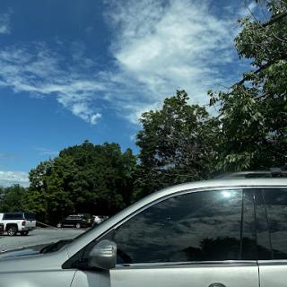 A silver car parked under a blue sky with fluffy clouds and green trees.