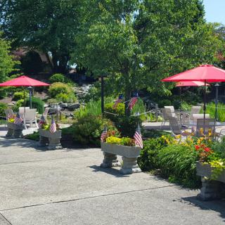 A sunny outdoor garden patio with red umbrellas, surrounded by lush greenery and colorful flowers.