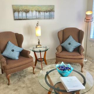 A cozy living room featuring two armchairs with decorative pillows, glass side and coffee tables, a table lamp, floor lamp, and wall artwork.