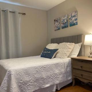 A cozy bedroom with a neatly made bed, featuring white bedding and decorative pillows, accompanied by a nightstand with a lamp and wall art above the headboard.