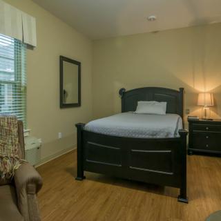 A cozy bedroom with a single bed, nightstand, and chair, featuring hardwood floors and soft lighting.