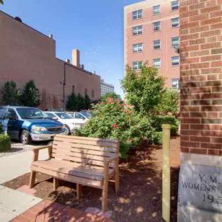 An urban scene featuring a wooden bench beside a brick wall, with parked cars and buildings in the background under a clear blue sky.