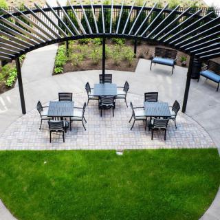 An outdoor patio with tables and chairs under a pergola, surrounded by a green lawn.