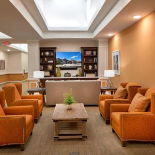 A modern lounge area featuring orange chairs, a central coffee table, and a bookshelf against one wall, all under bright lighting.