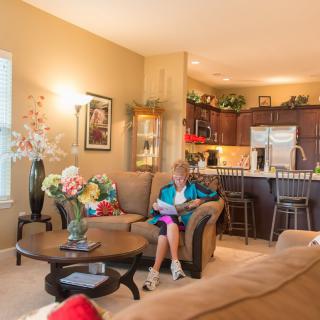 A cozy living room with a woman reading on a sofa, surrounded by decor and flowers, with an open view of the kitchen.