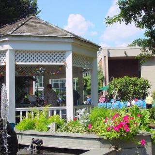 A picturesque gazebo surrounded by vibrant flowers and lush greenery near a small pond.