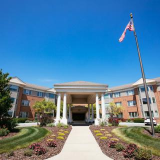A large building with a grand entrance, surrounded by landscaping, under a clear blue sky with a flagpole in front.