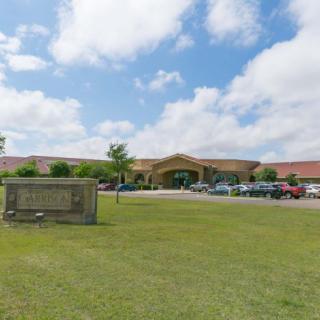 A school building with a parking lot, surrounded by grass under a partly cloudy sky.
