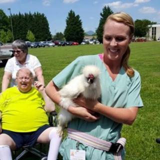 A nurse holding a fluffy dog stands next to an elderly person in a wheelchair, enjoying a sunny day outside.