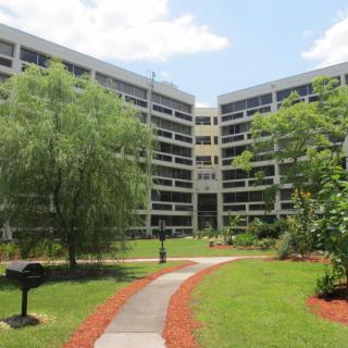 An outdoor area with a central pathway leading to a modern multi-story building, surrounded by lush greenery and trees under a partly cloudy sky.