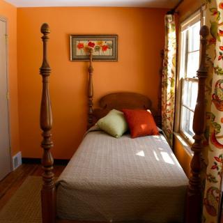 A cozy bedroom with orange walls features a twin bed adorned with colorful decorative pillows and floral curtains framing the window.
