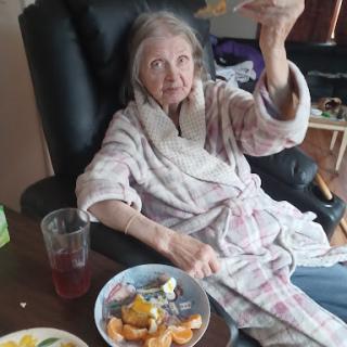 An elderly woman in a robe enjoying a snack while sitting comfortably.