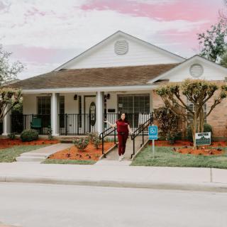 A person stands in front of a house with a porch, surrounded by landscaping and trees.