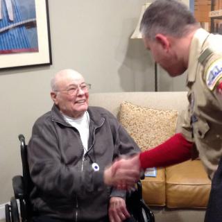 An elderly person in a wheelchair greets someone in a uniform with a handshake indoors.