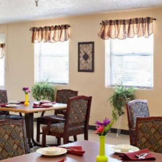 A well-lit dining area with neatly arranged tables and chairs, complemented by decorative plants and window treatments.