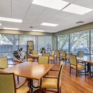 A bright and spacious room featuring dining tables, chairs, exercise equipment, and large windows.