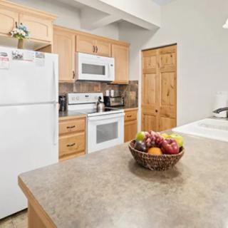 A modern kitchen with wooden cabinets, featuring a refrigerator, stove, microwave, and a fruit bowl on the countertop.