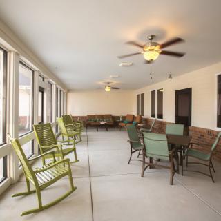 A spacious sunroom featuring rocking chairs, a dining table, and multiple windows with ceiling fans.
