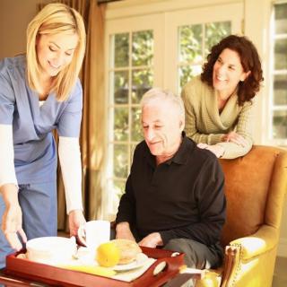 A caregiver assists an elderly man at home while a woman looks on.