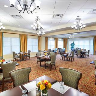 A spacious and elegantly decorated dining room with multiple tables and chandeliers, featuring large windows and a warm carpeted floor.