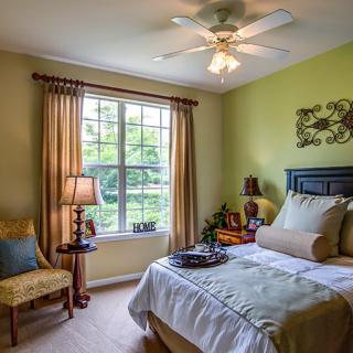 A cozy bedroom with a bed, chair, and lamp, illuminated by natural light from a large window.