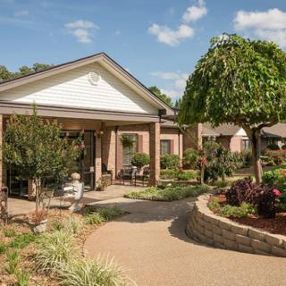 A charming outdoor garden area with a paved pathway leading to a small building, surrounded by lush greenery and manicured plants.