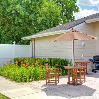 A sunny backyard patio with wooden furniture, an umbrella, and a barbecue grill surrounded by greenery.