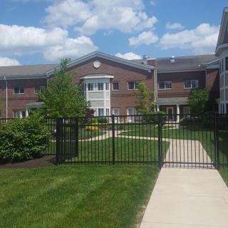A view of an apartment building with a fenced pathway and well-maintained grassy area under a partly cloudy sky.