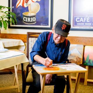 An artist wearing a cap is focused on painting while seated indoors.