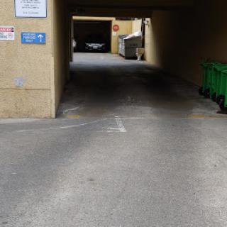 An urban alleyway leading to a parking area, with green garbage bins lined up on the right.