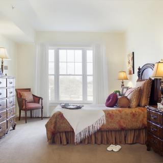 A cozy bedroom with classic furniture, featuring a bed, armchair, dressers, and a large window.