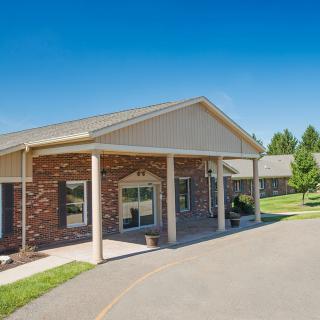 A one-story brick building featuring a covered entrance with columns, surrounded by a driveway and well-maintained lawn.
