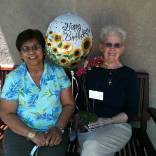 Two women sitting on a bench outdoors, celebrating a birthday with a balloon and flowers.