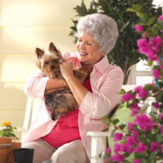 An elderly woman smiling and holding a small dog while surrounded by colorful flowers in a garden.