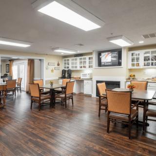 A well-lit dining area with wooden flooring, featuring tables, chairs, cabinets, and a television.