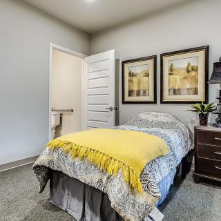 A cozy bedroom featuring a single bed with a yellow blanket, paintings on the wall, and a nightstand with a lamp.