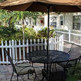 A cozy patio with a metal table and chairs under a large umbrella, surrounded by a white fence and lush tropical plants.