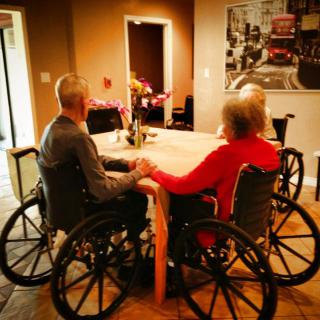 Elderly individuals sitting at a table in wheelchairs within a care home setting.
