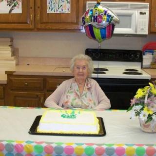 An elderly woman celebrating her birthday with a cake and balloon in a kitchen setting.