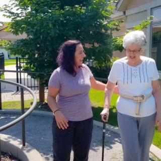A caregiver assisting an elderly woman walking outdoors on a sunny day.