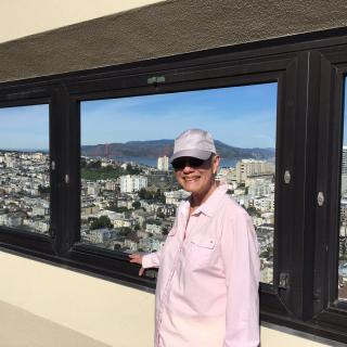 A person wearing a hat and sunglasses stands by a large window overlooking a cityscape.