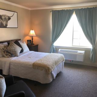 A cozy bedroom with a neatly made bed, soft lighting from a lamp, and large windows with blue curtains.