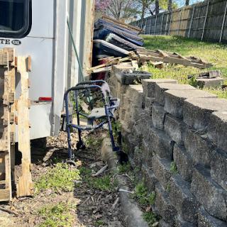 A wheelchair leaning against a concrete wall amidst clutter in a backyard.