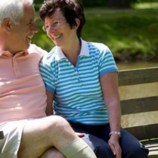 An elderly couple sitting on a bench, smiling at each other in a park.