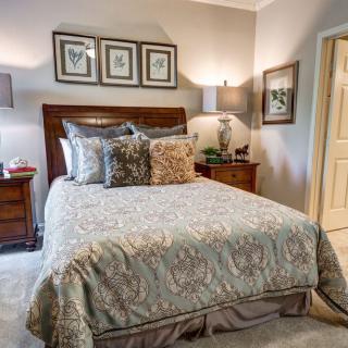 A cozy bedroom featuring a neatly made bed with multiple pillows, flanked by nightstands and lamps. The walls are adorned with framed artwork, creating a warm and inviting atmosphere.