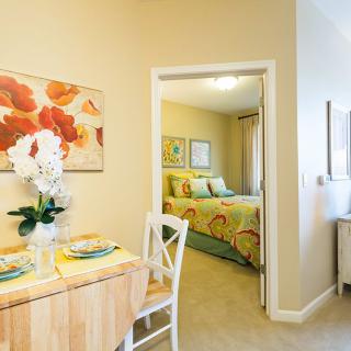A cozy apartment interior featuring a dining table with floral centerpiece, an adjoining bedroom with vibrant bedding, and decorative artwork on the walls.